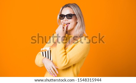 Young cheerful woman in 3D glasses watching a blockbuster and eating popcorn. Fan movie concept on yellow background, copy space
