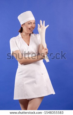 young woman doctor nurse in a white coat and hat with disposable gloves in her hands indulges and poses on a blue background