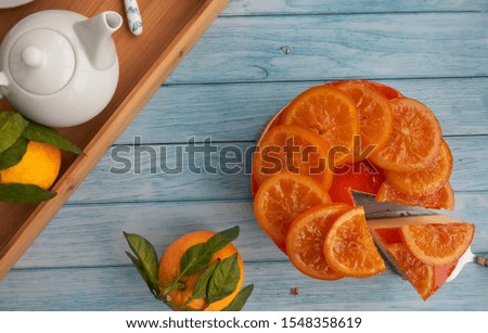 Top view and close-up of a cold homemade orange pie with candied orange on the surface, a piece of orange pie, fresh oranges with green leaves, a tray with a teapot on a blue background