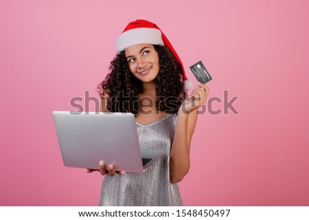 beautiful black woman with credit card and laptop wearing holiday santa hat and dress isolated over pink
