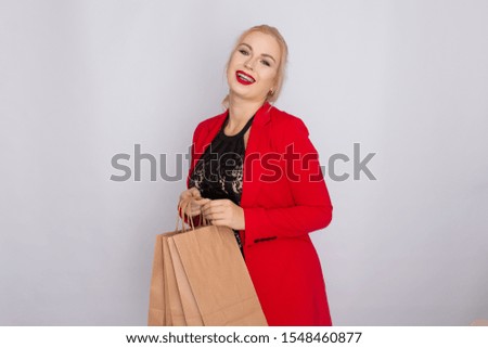 Image of excited blonde woman in red jacket holding brown shopping bags isolated over white bakcground