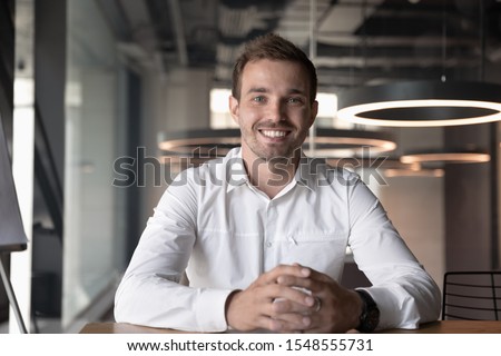Portrait of smiling male Caucasian employee sit at office desk look at camera in modern office, happy man worker millennial businessman posing, having video call with client or customer online