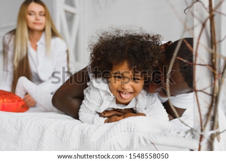 Portrait of happy multiracial young family lying on cozy white bed at home, smiling international mom and dad relaxing with little biracial girl child posing for picture in bedroom