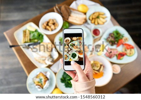 A blogger takes a photo on a smartphone for social networks. Table with Plates seafood BBQ with garnish. Variety of dishes on the table. Restaurant menu.