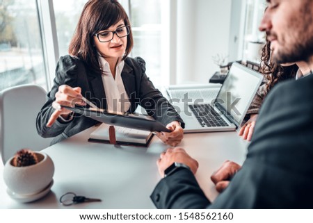 Mature female estate agent showing terms of agreement in contract to young family couple sitting at the desk in bright office. Dealership, home purchasing, signing a contract concept