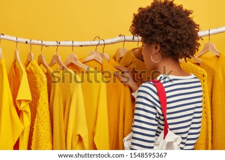 Back view of curly woman in sailor jumper, carries bag, selects clothes on racks, chooses outfit for future important event, chooses yellow cape on hangers, makes purchasing in fashion store