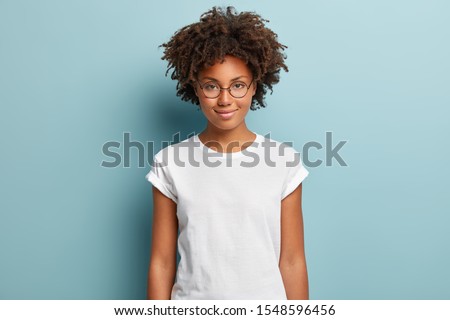 Attractive female student with curly hair, wears transparent glasses, white t shirt, stands against blue background, has calm face expression, tender smile, listens interlocutor with pleasure Royalty-Free Stock Photo #1548596456