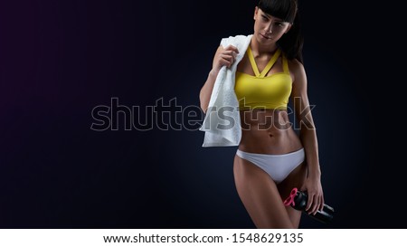 Close-up photo of female with perfect abdomen muscles on dark background with copyspace. Protein shake bottle and white towel in her hands.