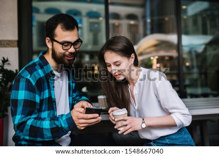 Positive multiracial male and female friends watching photos on smartphone spending coffee break together holding to go cups, smiling students hipsters checking videos on cellular from social networks