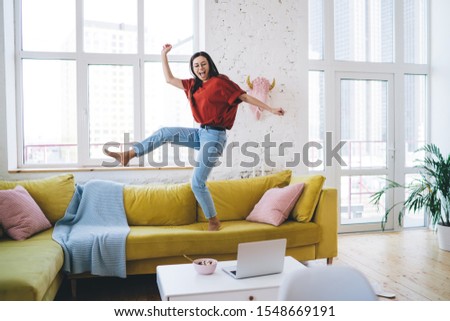 Young smiling woman in casual clothing and glasses dancing on yellow couch in living room near coffee table with laptop and breakfast bowl Royalty-Free Stock Photo #1548669191