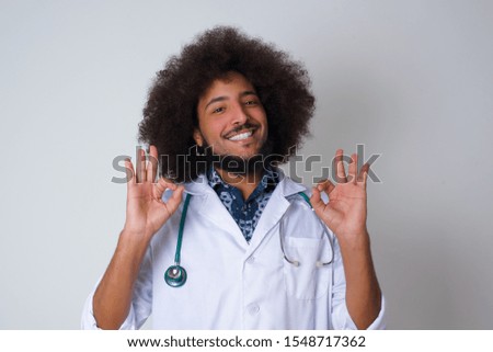 Glad attractive African American doctor man shows ok sign with both hands as expresses approval, has cheerful expression being optimistic. Standing against gray wall.