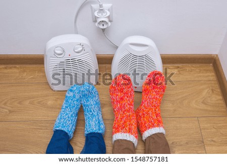 Family dressed in warm knitted woolen orange and blue socks near an electric fan heaters in cold winter time. The symbolic image of the heating season at home. Part of body, selective focus.