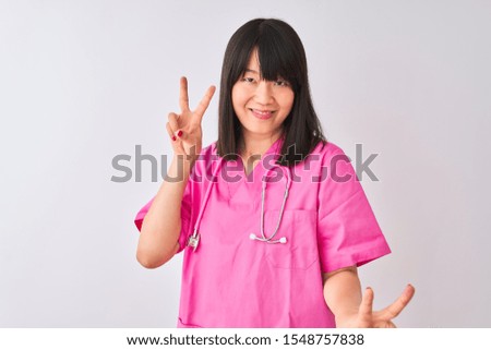 Young beautiful Chinese nurse woman wearing stethoscope over isolated white background smiling looking to the camera showing fingers doing victory sign. Number two.