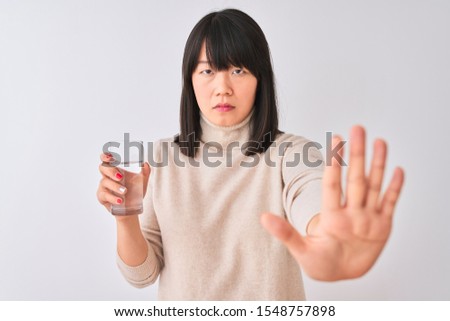 Young beautiful Chinese woman holding glass of water over isolated white background with open hand doing stop sign with serious and confident expression, defense gesture