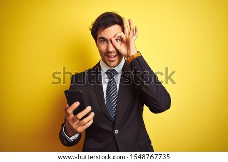 Young handsome businessman using smartphone standing over isolated yellow background with happy face smiling doing ok sign with hand on eye looking through fingers