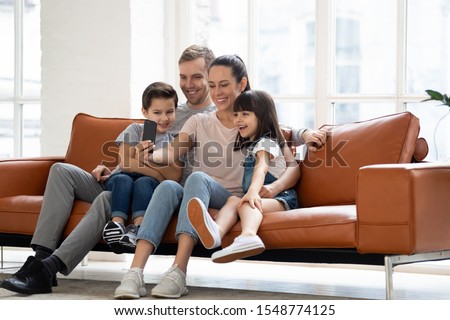 Happy young family with two kids sit on couch making self-portrait picture on smartphone together, smiling parents and children have video call use fast unlimited wireless internet connection on cell