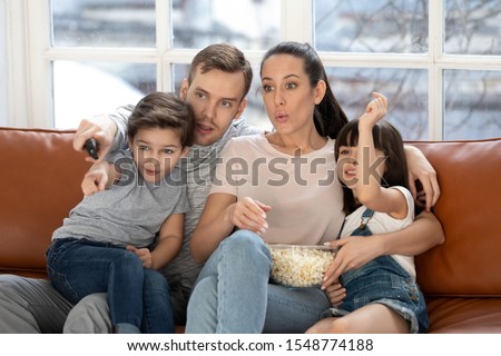 Young parents sit on couch relax with cute little kids watching movie or cartoon on tv together, mom and dad rest at home enjoy video eating popcorn with preschooler children, family weekend concept