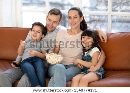 Smiling young family with preschooler children sit relax on sofa watching movie on tv eating popcorn, happy parents rest at home with little kids enjoy weekend together, have fun in living room