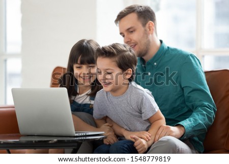 Excited small preschooler boy and girl kids have fun watching cartoon on laptop with young caring dad, loving father relax spend time with little children using computer, play game online or learning