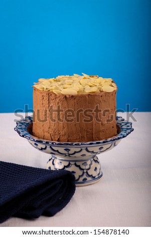 Chocolate birthday party cake with almond flakes on antique ceramic stand with pattern, blue background, white canvas