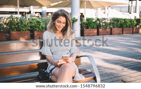 Attractive Caucasian hipster photographer with modern camera resting on bench and searching famous showplaces of Barcelona for taking pictures on leisure connected to 4g wireless on cellphone