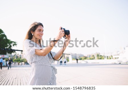 Positive female photographer in stylish dress focusing for clicking good pictures of Barcelona landscape, happy Caucasian woman tourist with modern camera taking pistures during Spanish vacations