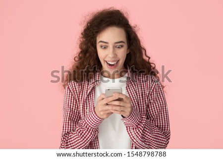 Overjoyed millennial girl with curly hair wearing casual shirt isolated on pink studio background read pleasant good news on cellphone, happy young female excited by unexpected great message on cell