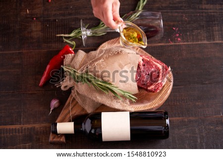 hand, with a container of olive oil, next to a piece of juicy, seasoned ribeye steak wrapped in paper and burlap on a cutting board on a wooden background. Nearby is a bottle of red wine, a wine glass