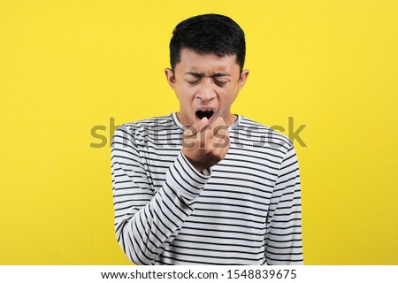 Portrait of funny Asian man yawning covering open mouth and showing a sleepy gesture. feeling being tired from hard work, isolated on yellow background