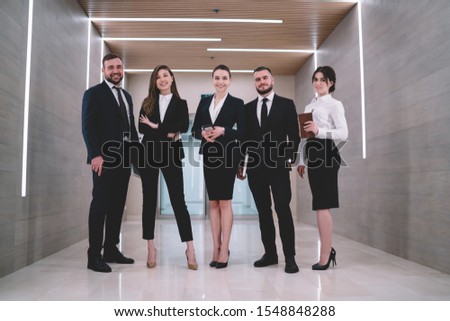 Group of successful and presentable colleagues in formal suits standing in bright corridor decorated by creative lamps and looking at camera Royalty-Free Stock Photo #1548848288