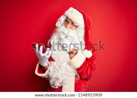 Middle age man wearing Santa Claus costume using tablet over isolated red background serious face thinking about question, very confused idea