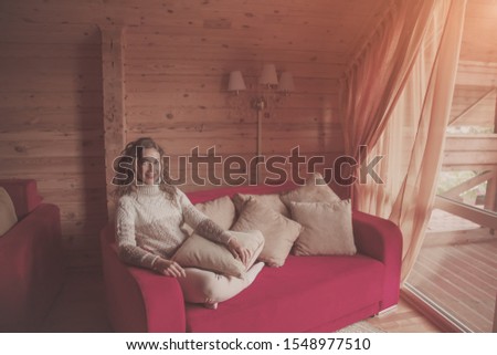Happy woman relax on red sofa in log house and look to the village nature in window. Woman dressed knitted sweater