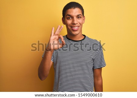 Young handsome arab man wearing navy striped t-shirt over isolated yellow background smiling positive doing ok sign with hand and fingers. Successful expression.