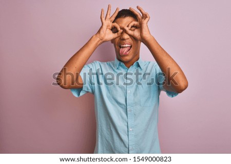 Young handsome arab man wearing blue shirt standing over isolated pink background doing ok gesture like binoculars sticking tongue out, eyes looking through fingers. Crazy expression.