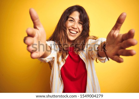 Young beautiful woman wearing red t-shirt and stripes shirt over yellow isolated background looking at the camera smiling with open arms for hug. Cheerful expression embracing happiness.