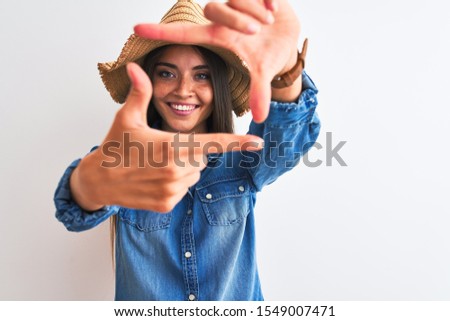 Young beautiful woman wearing denim shirt and hat standing over isolated white background smiling making frame with hands and fingers with happy face. Creativity and photography concept.