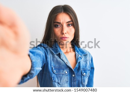 Beautiful woman wearing denim shirt make selfie by camera over isolated white background Relaxed with serious expression on face. Simple and natural looking at the camera.