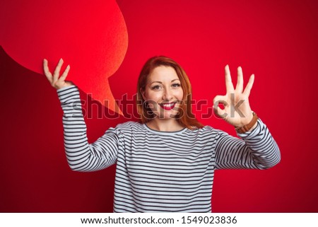 Young beautiful redhead woman holding speech bubble over red isolated background doing ok sign with fingers, excellent symbol