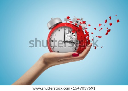 Side closeup of woman's hand facing up and holding red retro alarm clock that is dissolving into small pieces on light-blue background. Run out of time. Fail deadline. Stress out.