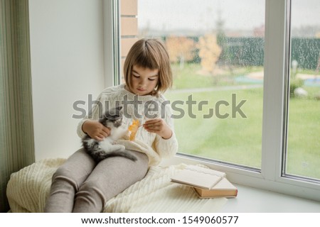 cute little girl sitting near the window, reading a book and holding a gray kitten in her arms Royalty-Free Stock Photo #1549060577