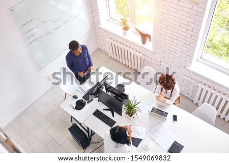 International partners exchange experience of working in business. Top view on three woman discussing, african coach stand speaking to others. white interior of office