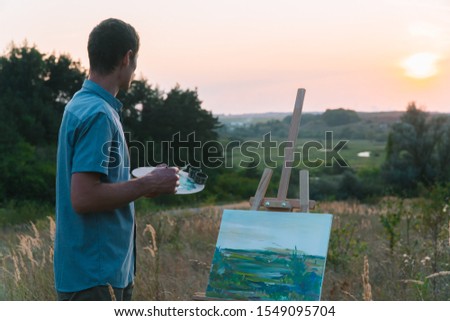 Male artist paints sunset landscape outdoors in nature landscape. A man paints a picture