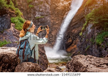 Happy traveller woman taking photo of majestic waterfall in the jungle. Hiking and sightseeing concept