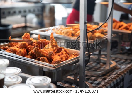 Stainless steel trays of fried chicken wings and other food sits underneath heat lamps at a buffet on a checkered table cloth. Royalty-Free Stock Photo #1549120871