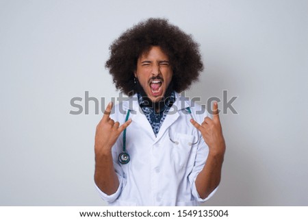 Born to rock this world. Joyful African American doctor man in medical uniform screaming out loud and showing with raised arms horns or rock gesture, expressing excitement of being on concert of band.