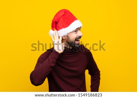 Man with christmas hat over isolated yellow background listening to something by putting hand on the ear