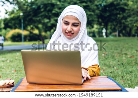 Portrait of happy arabic muslim woman with hijab dress smiling and using laptop cumputer in summer park