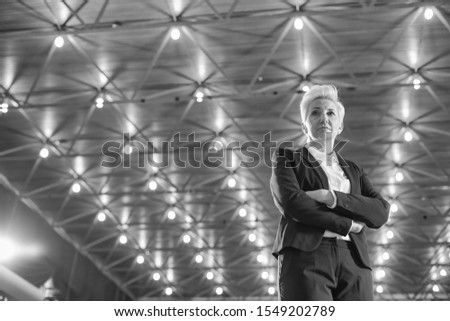 Black and white photo of Confident blond businesswoman standing with arms crossed against illuminated roof
