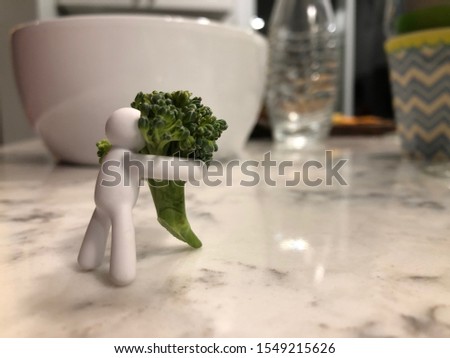 Little plain white figure hugging broccoli in the a kitchen, with a bowl, glass and water bottle in the background.