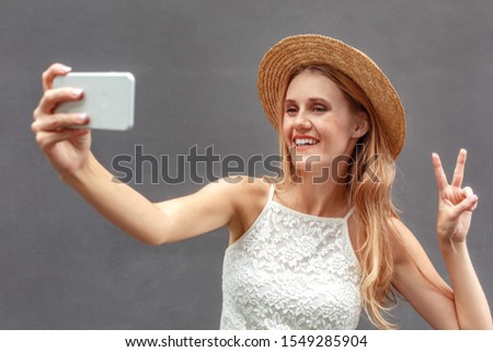 Young girl wearing hat standing isolated on wall taking selfie picture on smartphone posing to camera showing peace sign smiling playful close-up